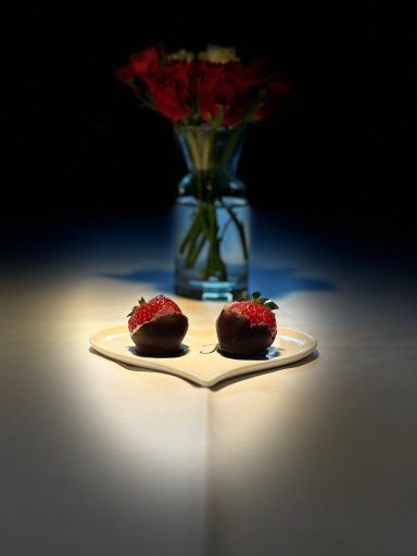 Zwei Schokoladeneerdbeeren vor einer Vase mit roten Blumen auf einem Tisch.